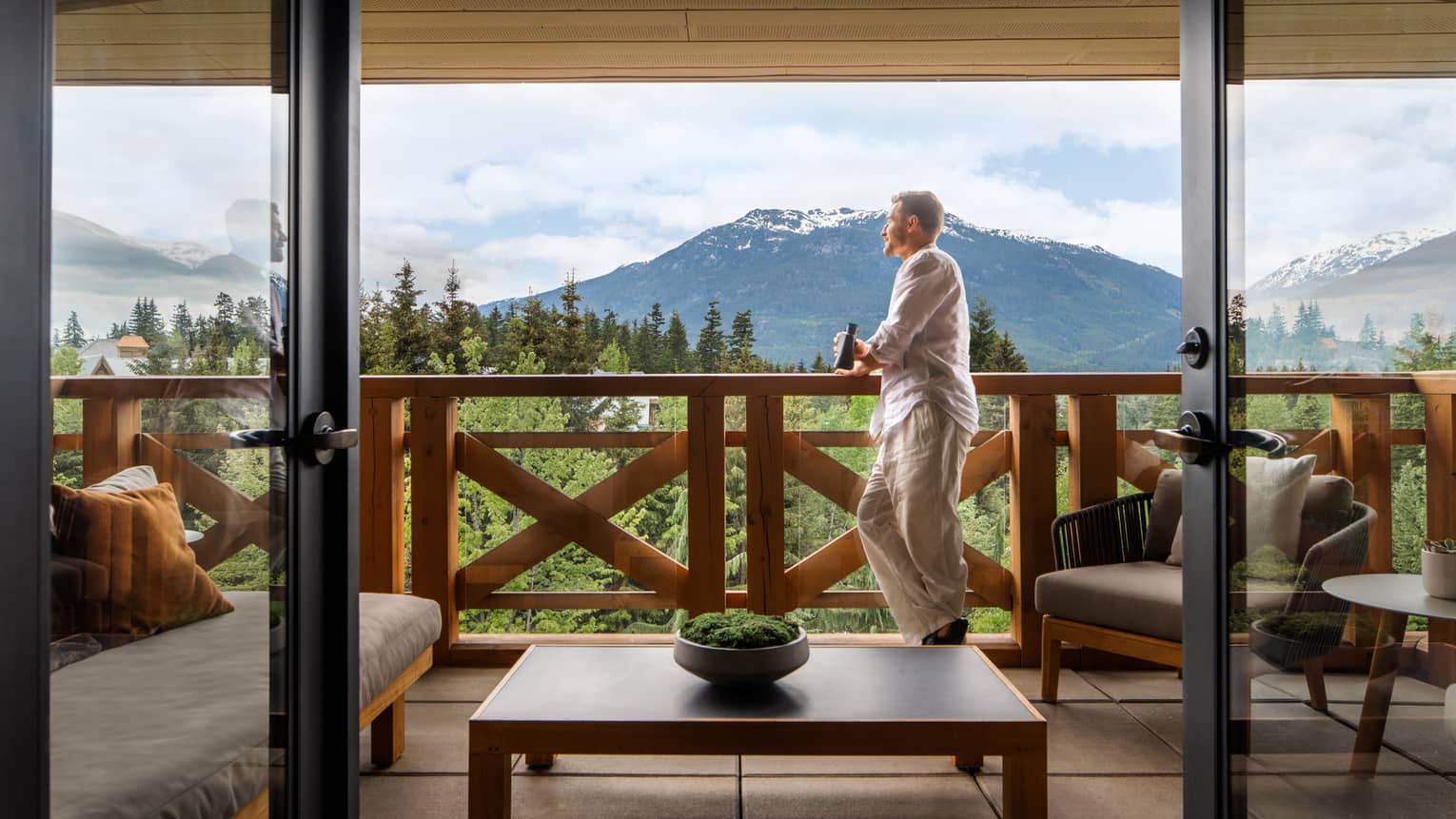 Man with short hair wearing a white long-sleeve shirt and khaki pants leans on the railing of a balcony with a beer in hand, looking out onto the mountain views