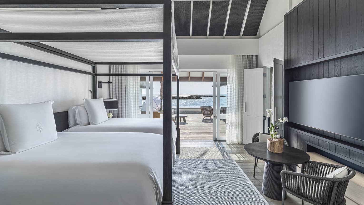 Bedroom with double four-poster canopied beds, wall-mounted flat-screen TV, two-seater round table, doorway opening onto deck with pool and cabana