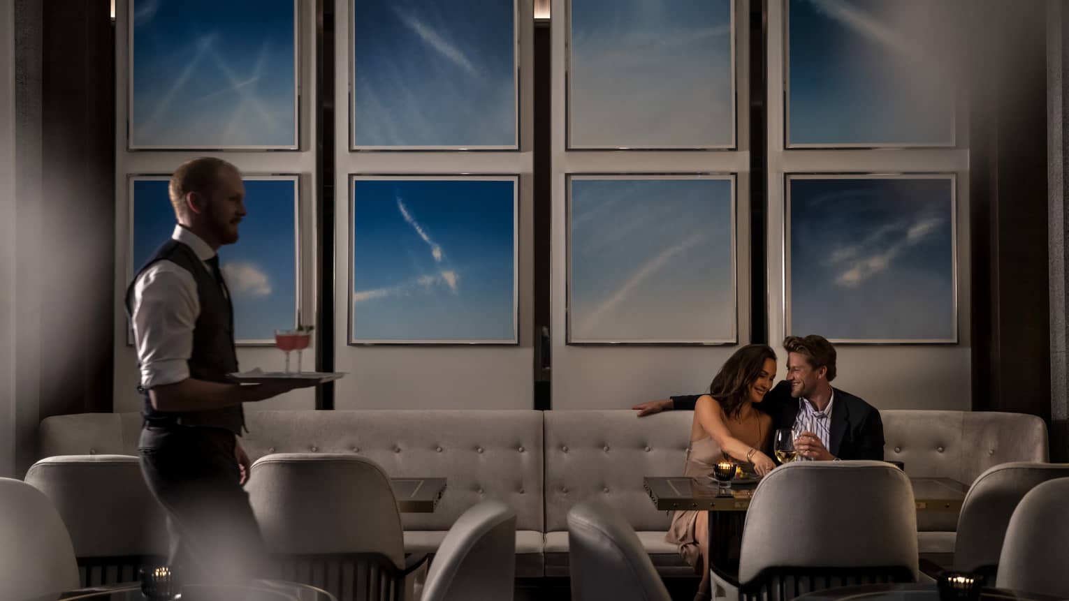 A bartender carrying two cocktails on a tray while a couple sits at a booth in the background of a restaurant.