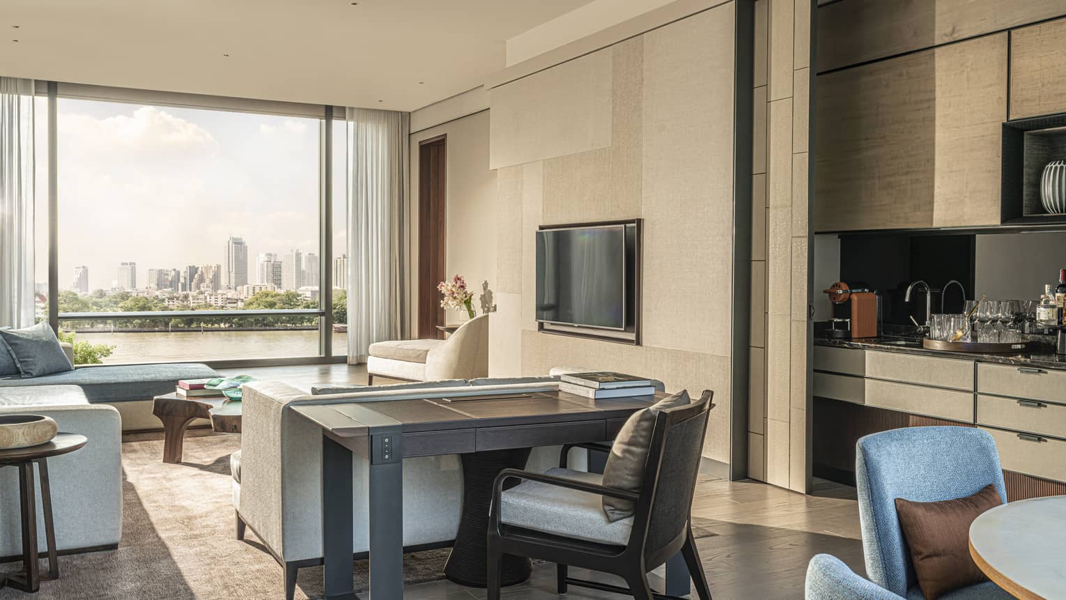 Hotel suite living area with blue, grey and beige furniture, console table, TV inset on beige wall, city view out large window
