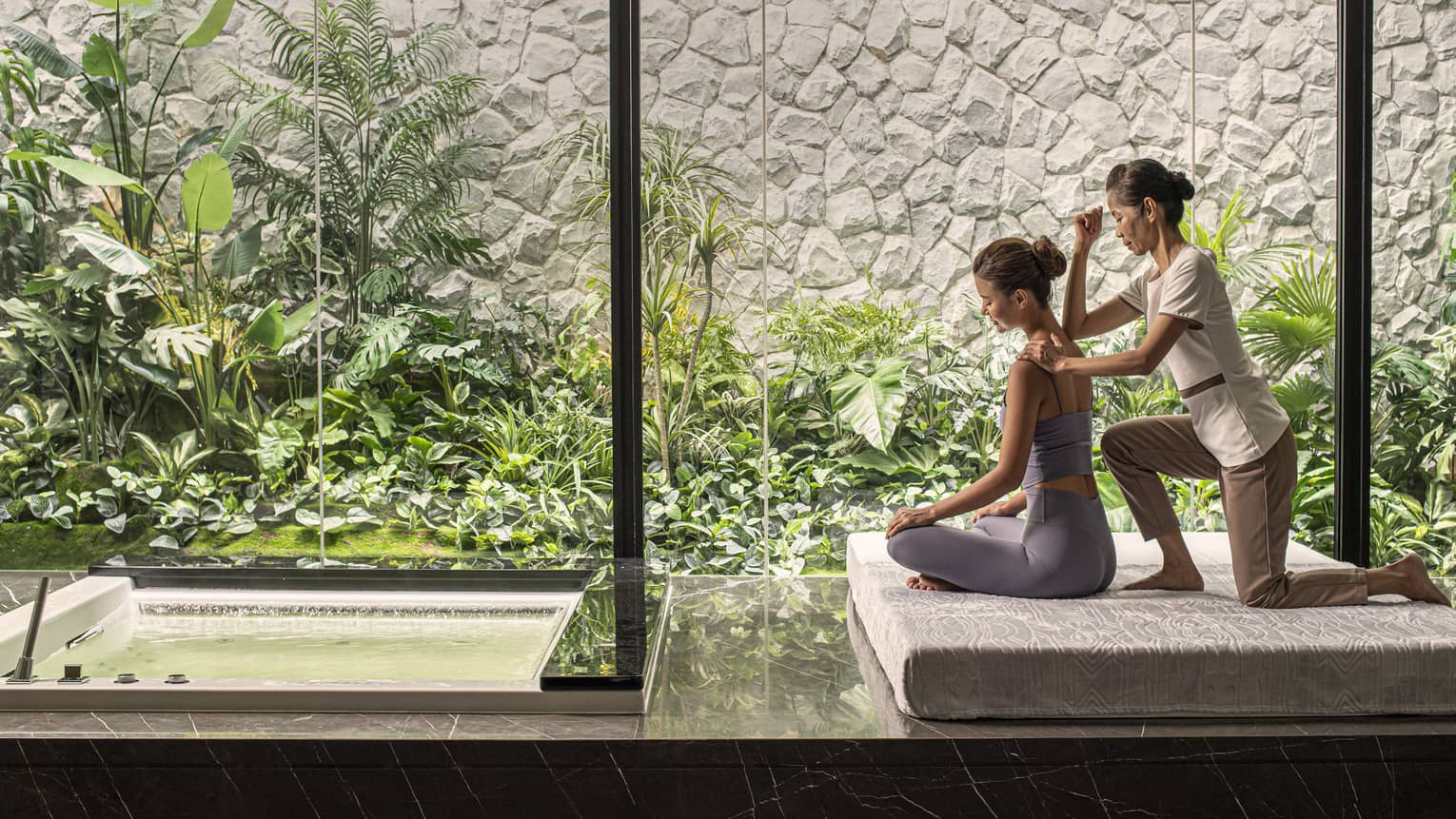 Person receiving a massage in a peaceful spa setting with large windows overlooking lush green plants and a small water feature.