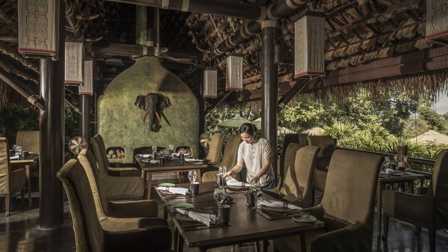 A woman sets the table in an open-air restaurant, natural-coloured furnishings, elephant head decoration on wall