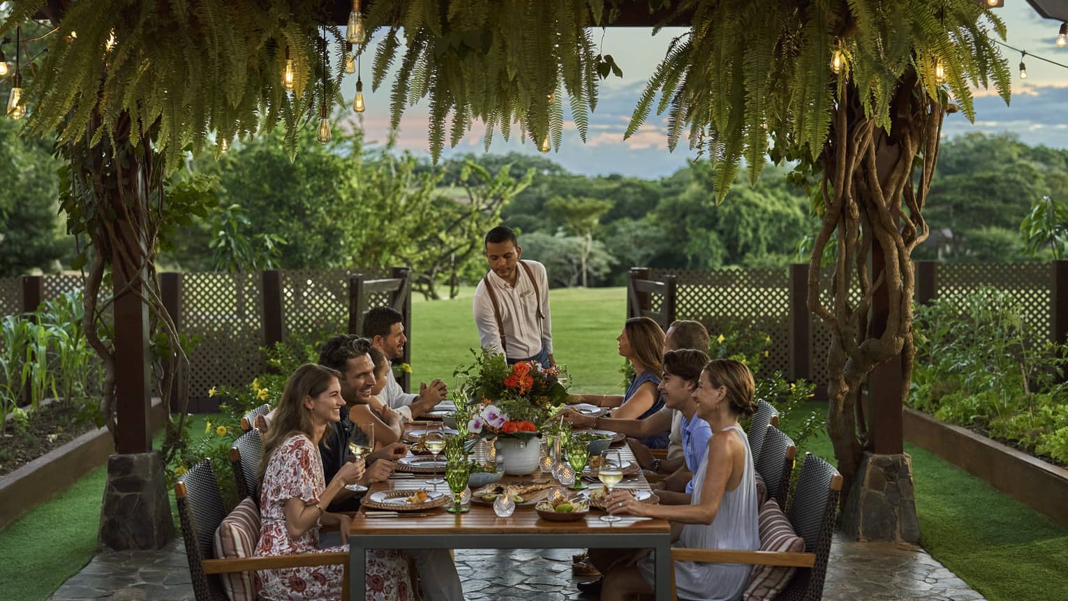 A group of people settled around a table, underneath a series of string lights and palm trees.