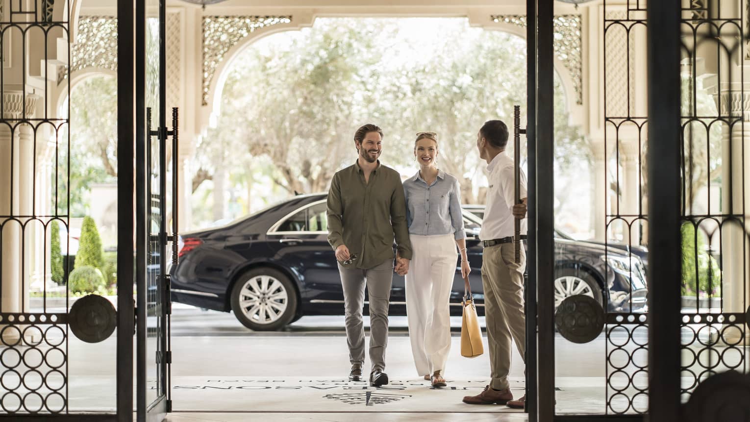 A well-dressed couple enters through the wrought iron doors of the hotel with a large blooming tree behind them.