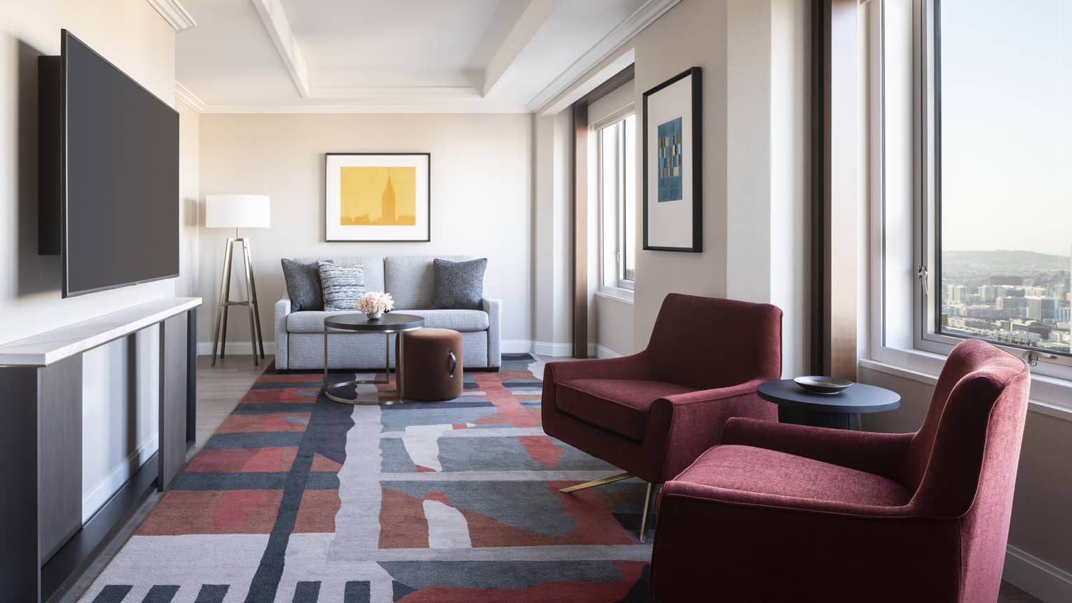 Hotel living room with two maroon arm chairs, carpet with geometric patterns, sofa, TV, windows