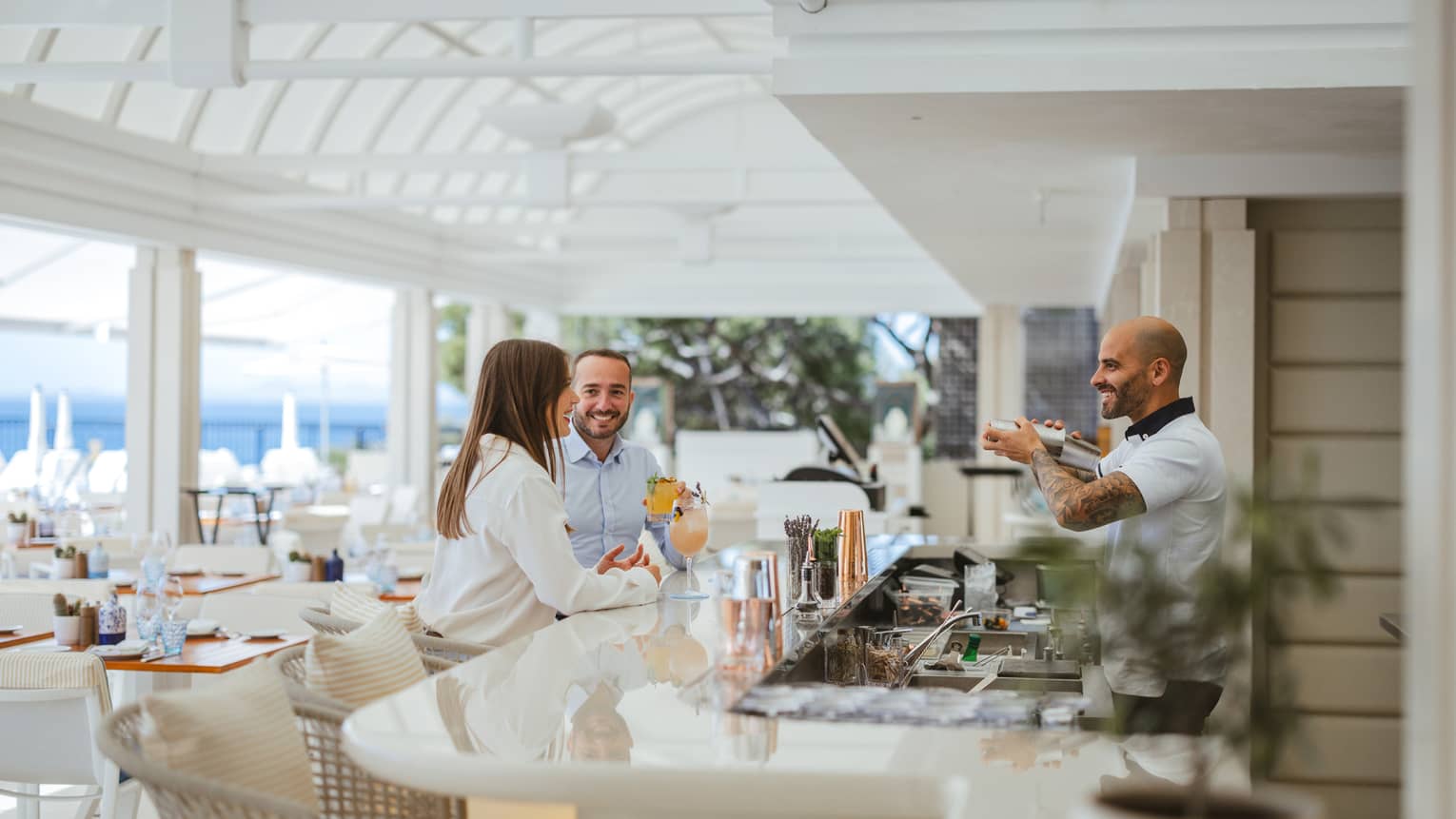Couple sitting at a white bar, talking with bartender, on restaurant?s open-air covered terrace