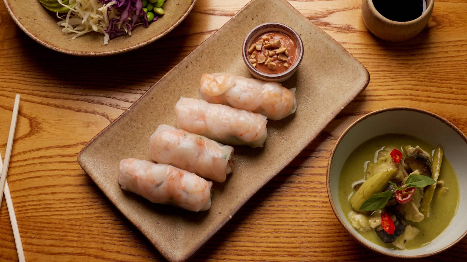 Row of shrimp rolls and sauce bowl on brown rectangular plate between fresh and pickled vegetable garnishes