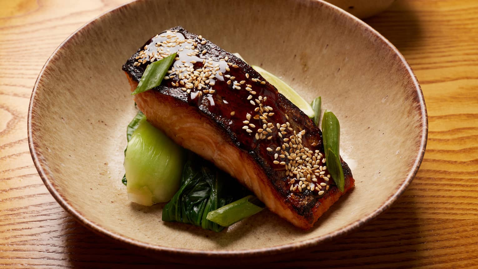 Seared sesame salmon garnished with sliced scallions atop bed of wilted bok choy in brown bowl