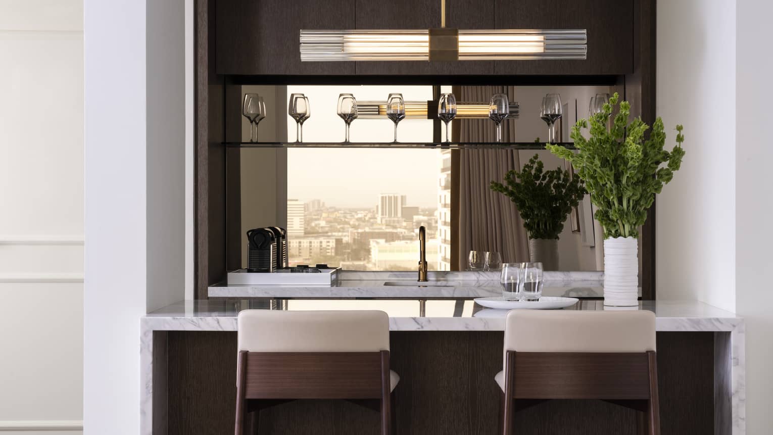 A kitchen with wine glasses on a shelf, herbs, two bar stools, an island and sink.