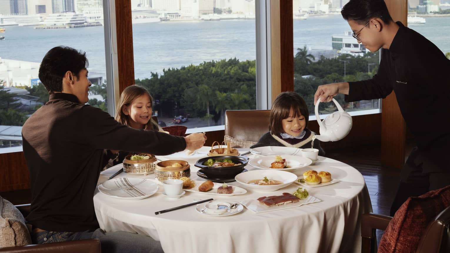 A hotel restaurant waiter pour tea at a white tablecloth table with man and two children, set next to a window overlooking city and harbour