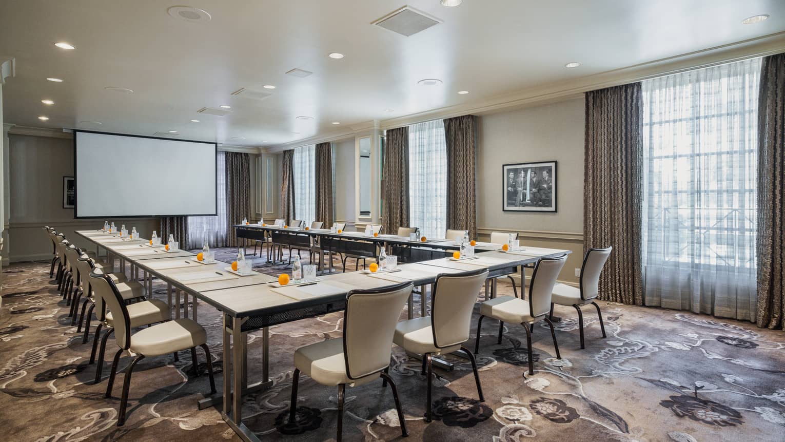 Meeting room set up hollow square style with projection screen, floor-to-ceiling windows and dark patterned carpet