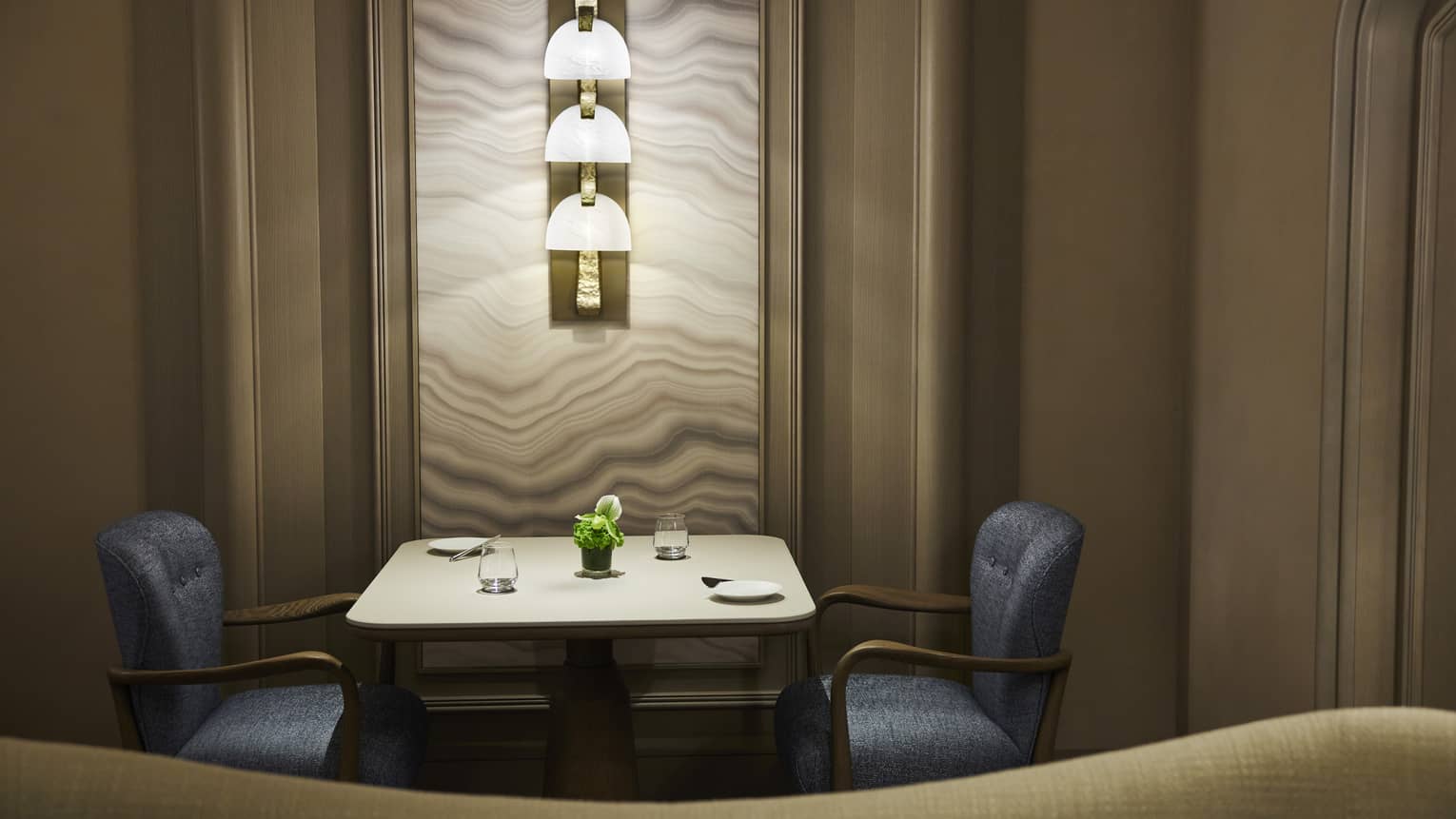 Dimly lit dining section with table set for two, lighted wall sconce above table, blue tufted chairs, tan colour scheme
