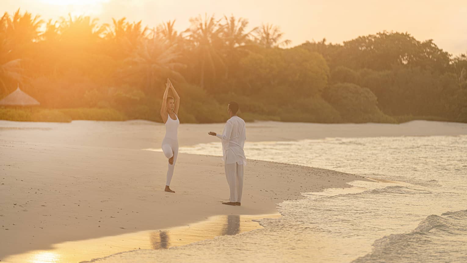 Person practicing yoga on a tropical beach at sunrise with an instructor, surrounded by calm ocean waves and lush greenery in the background