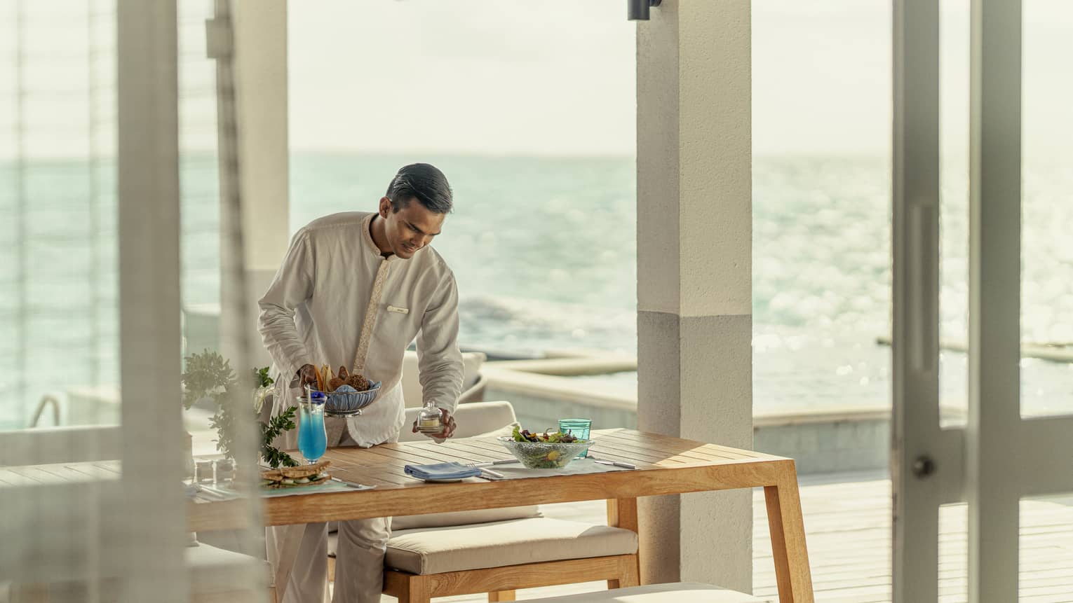 Male staff member sets table for In-Villa Dining, ocean in background
