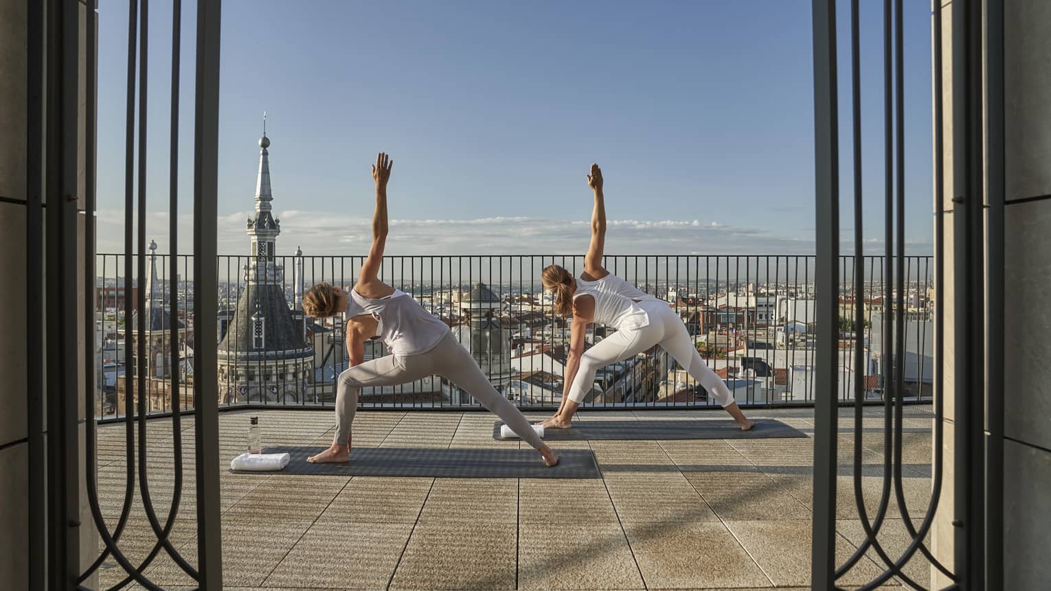 Two woman do triangle pose on outdoor terrace overlooking Madrid