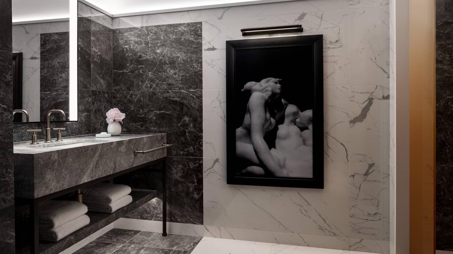 A sleek bathroom featuring dark marble walls and flooring, a large backlit mirror and a minimalist vanity with folded towels. A black-and-white art piece depicting sculptures hangs on the wall.