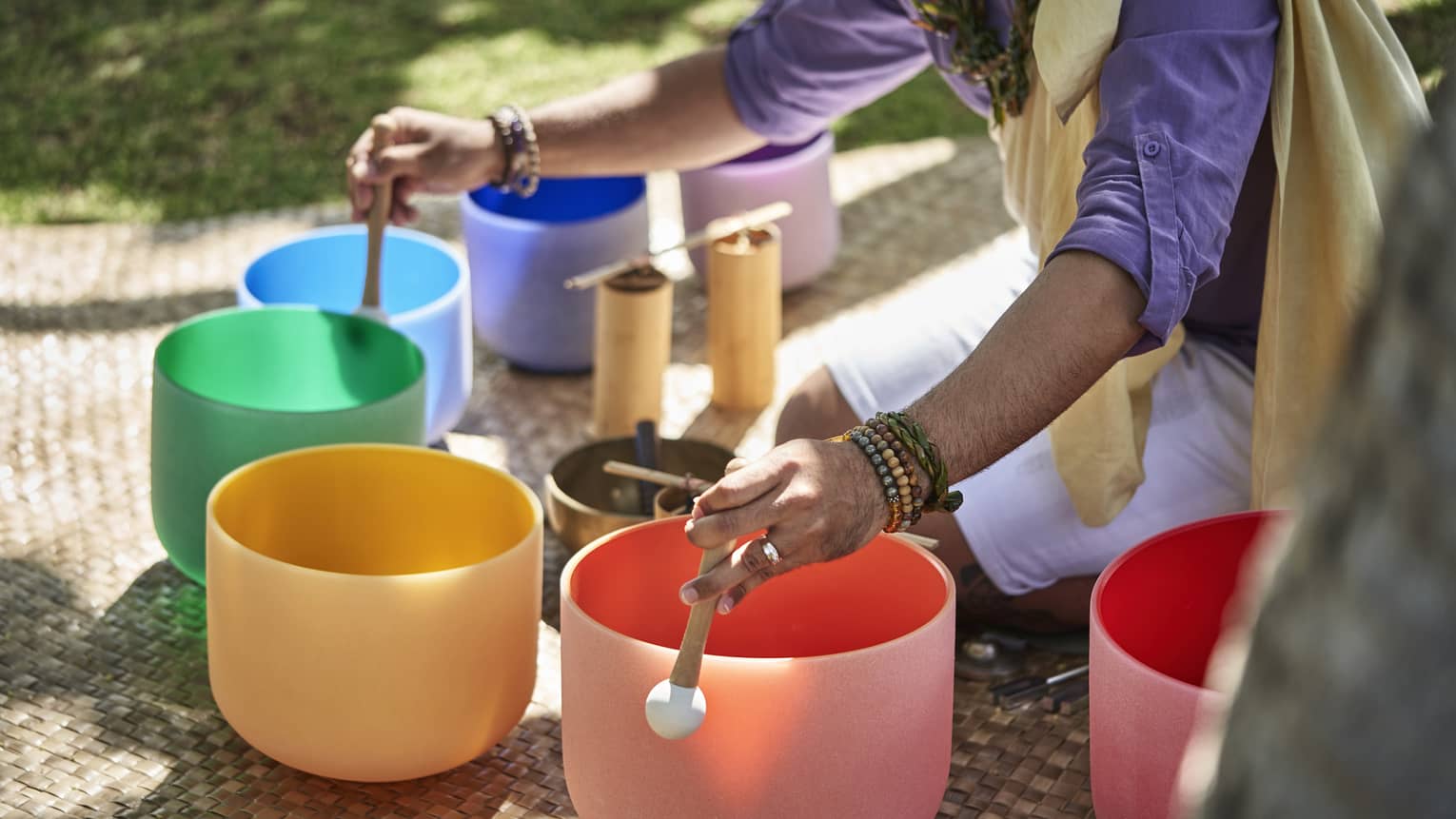 Close up of a man's hands using mallets to play a row of colourful crystal sound bowls
