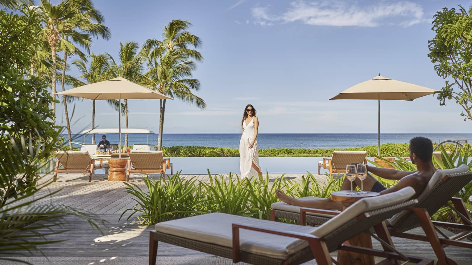 A woman dressed in white walks toward a lounge chair by an outdoor infinity pool