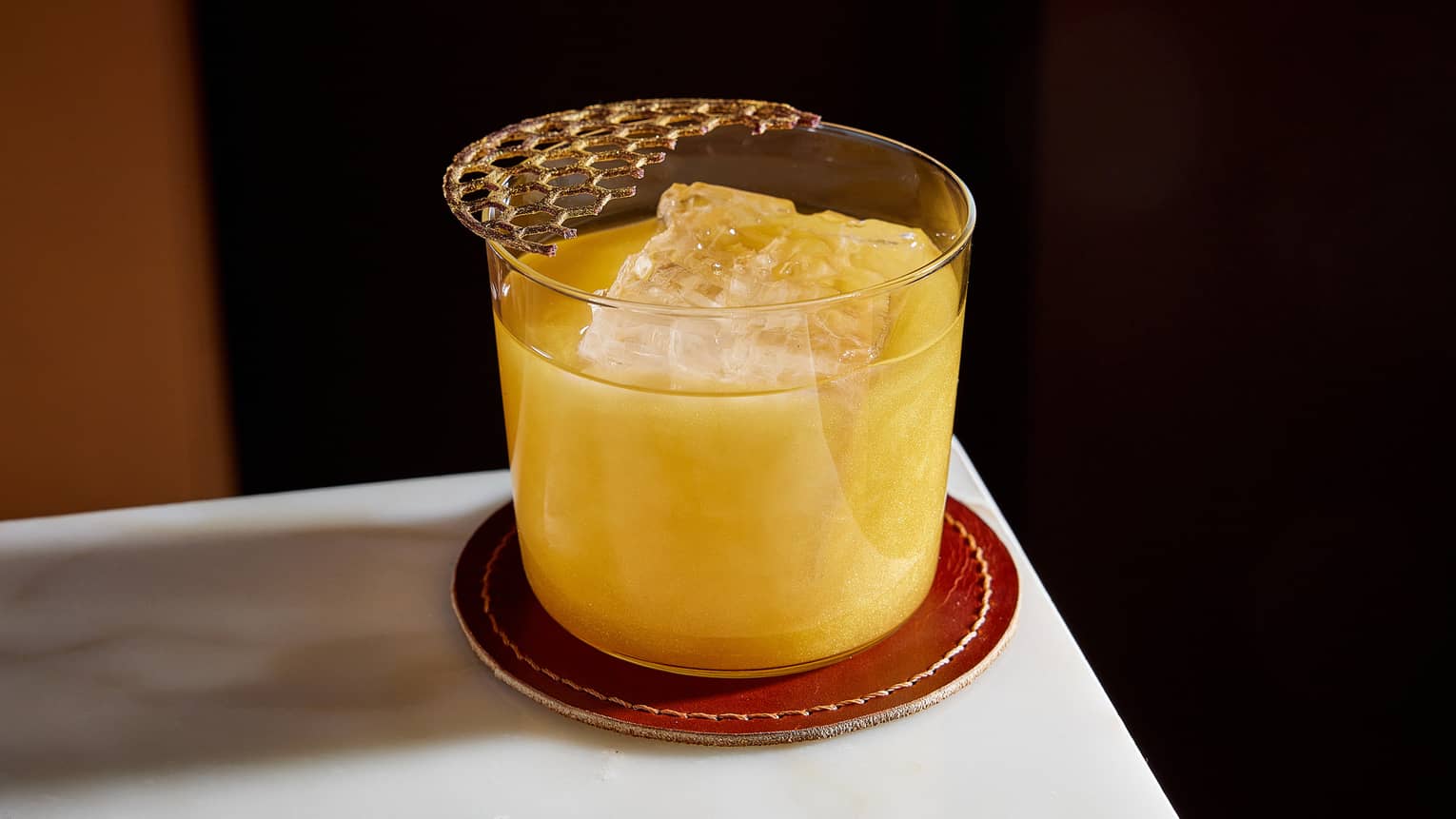 Golden cocktail on ice in old fashioned glass on leather coaster