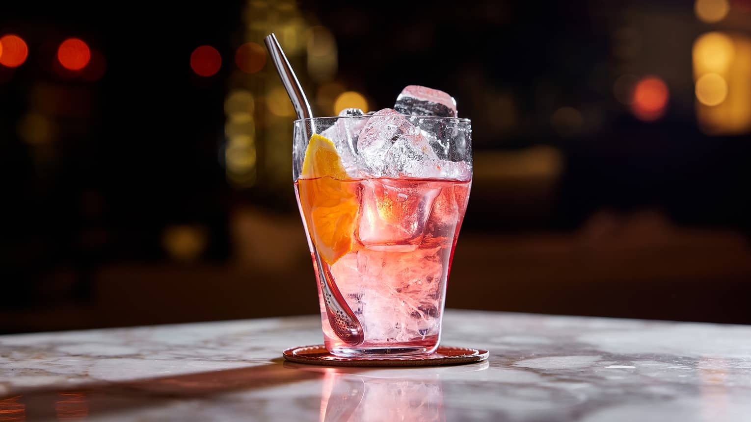 Pinkish-red cocktail with steel straw in highball glass filled with ice cubes