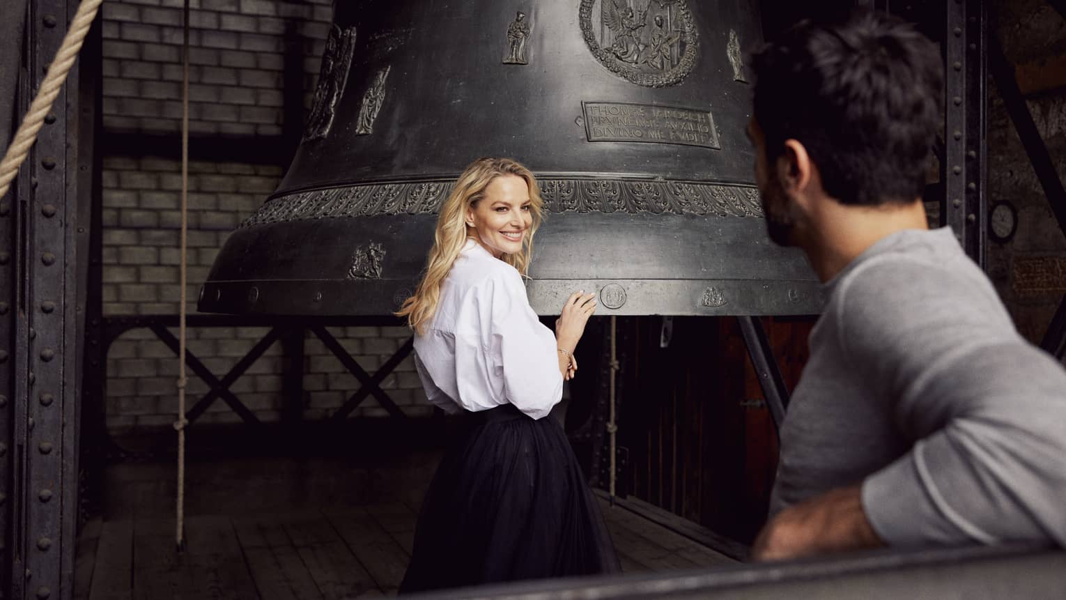 Person in white blouse standing if front of a large bell, smiling at another person