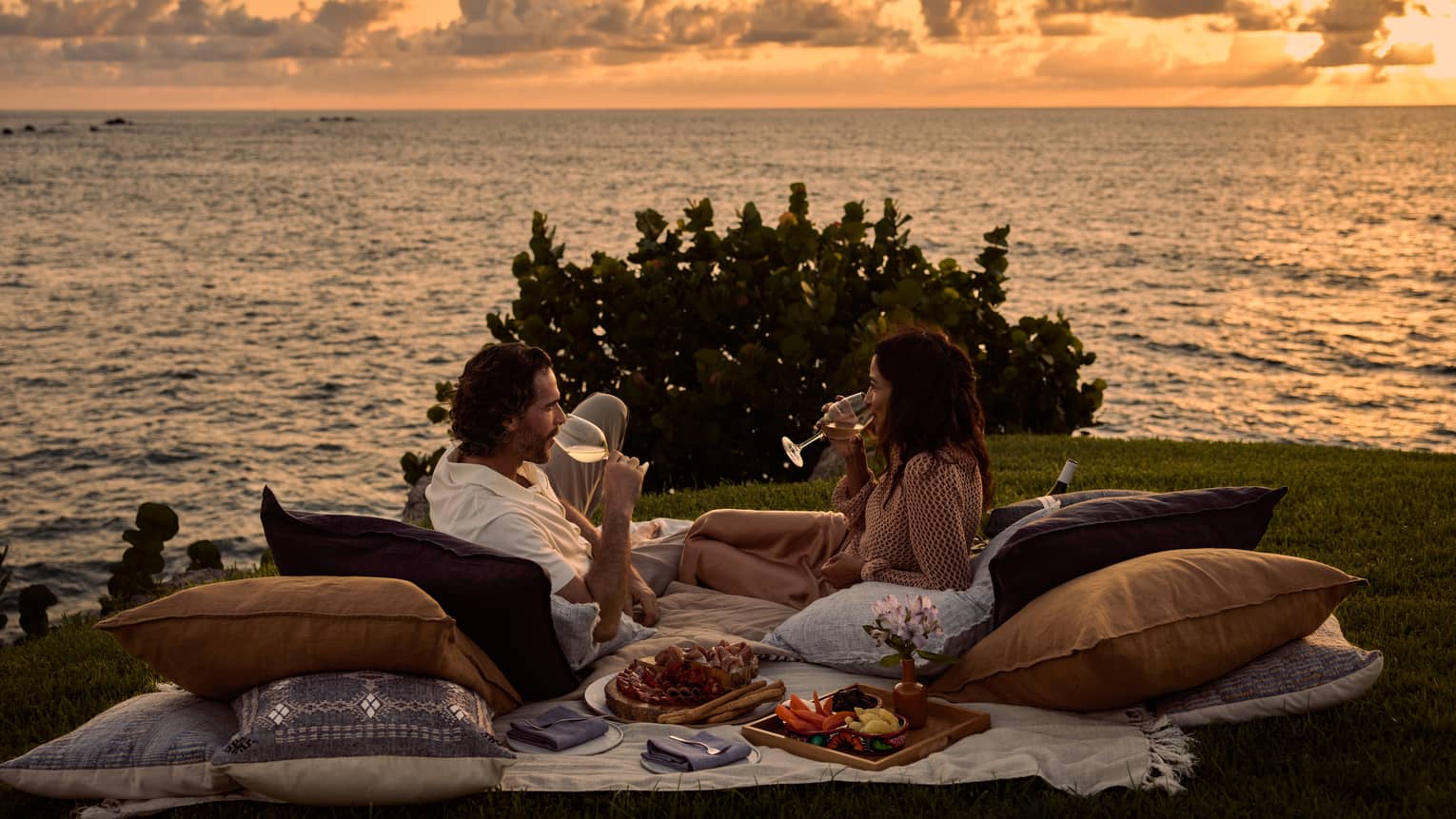 A man and woman lay on blanket and pillows during luxury picnic at sunset, next to the ocean
