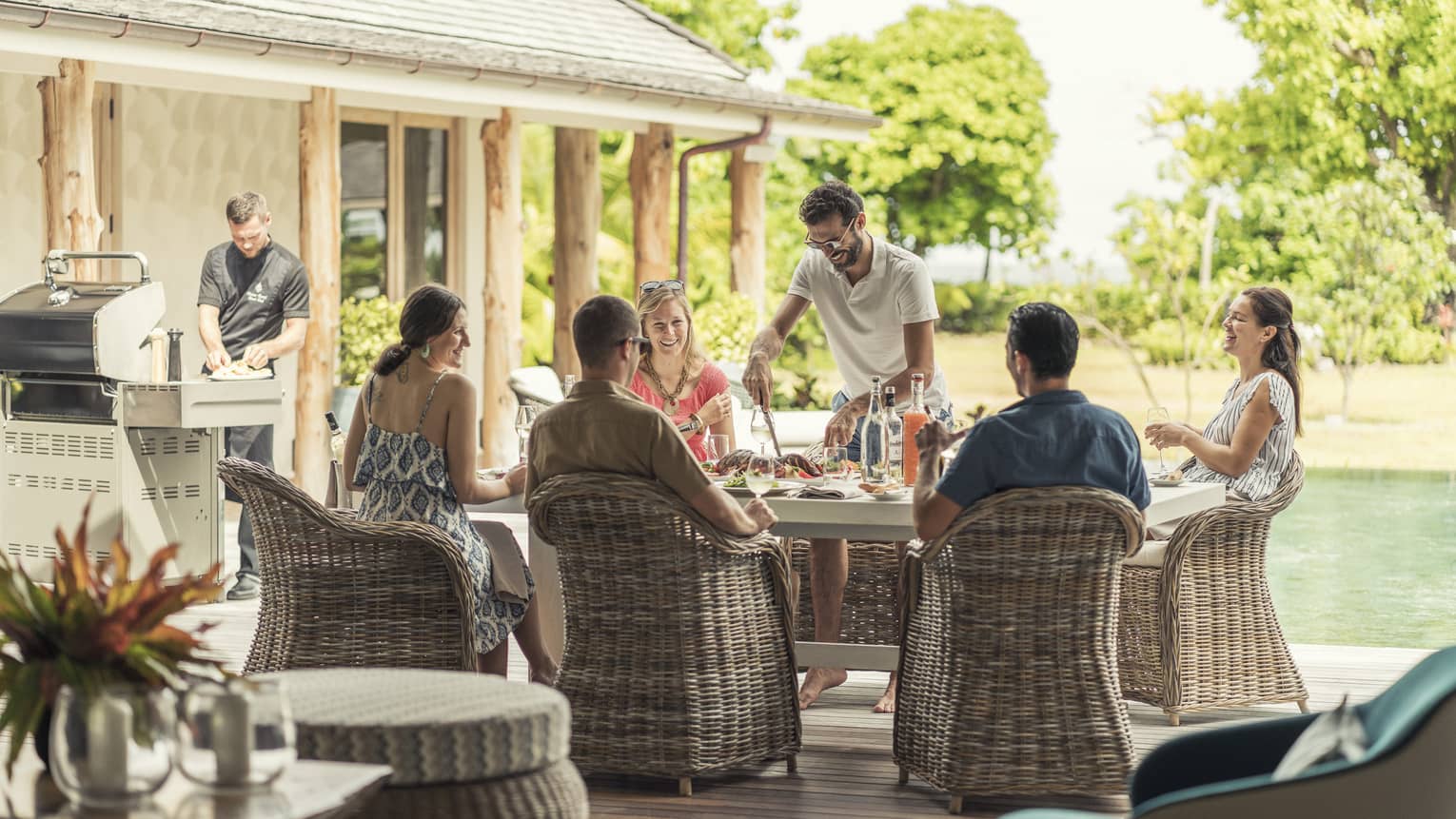 Group of friends enjoying an outdoor meal together on a sunny patio, with a chef preparing food on a grill nearby, creating a relaxed and social atmosphere.