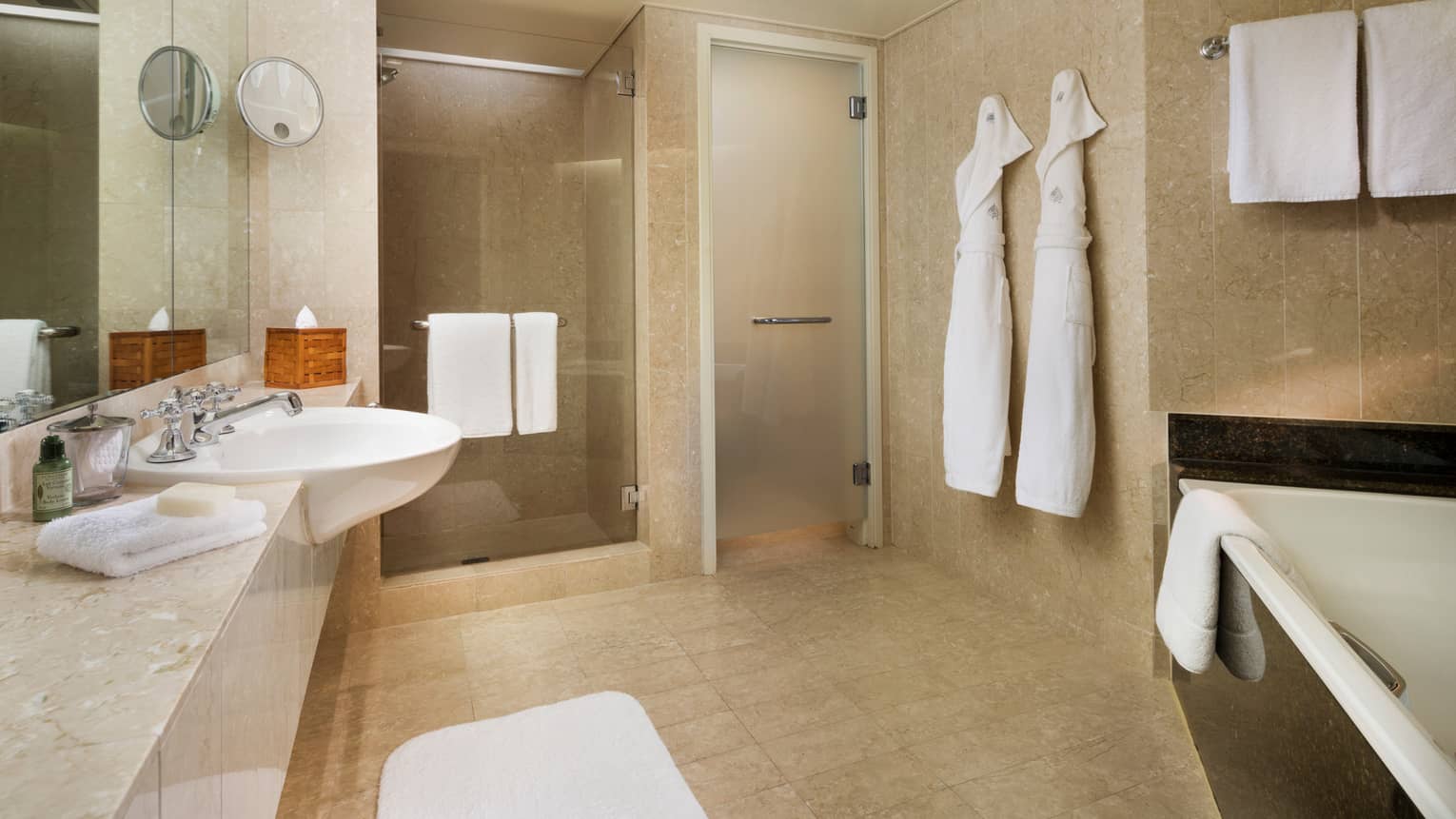 Hotel Suite full marble bathroom, two white robes hang by glass walk-in showers, sink