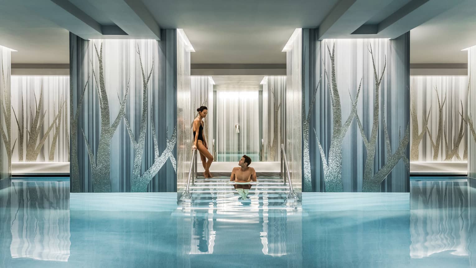A couple lounges on the steps of a luxurious, serene indoor pool flanked by wide pillars decorated with a sparkly tree motif.