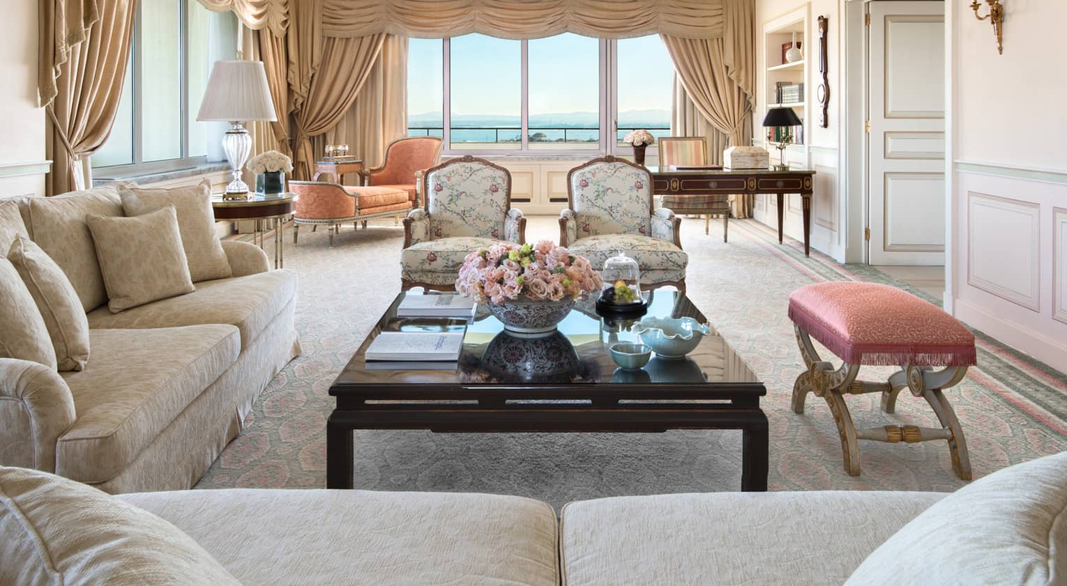 Elegant living room with cream sofas, floral armchairs, a dark wood coffee table, ornate drapes and large windows with a water view.