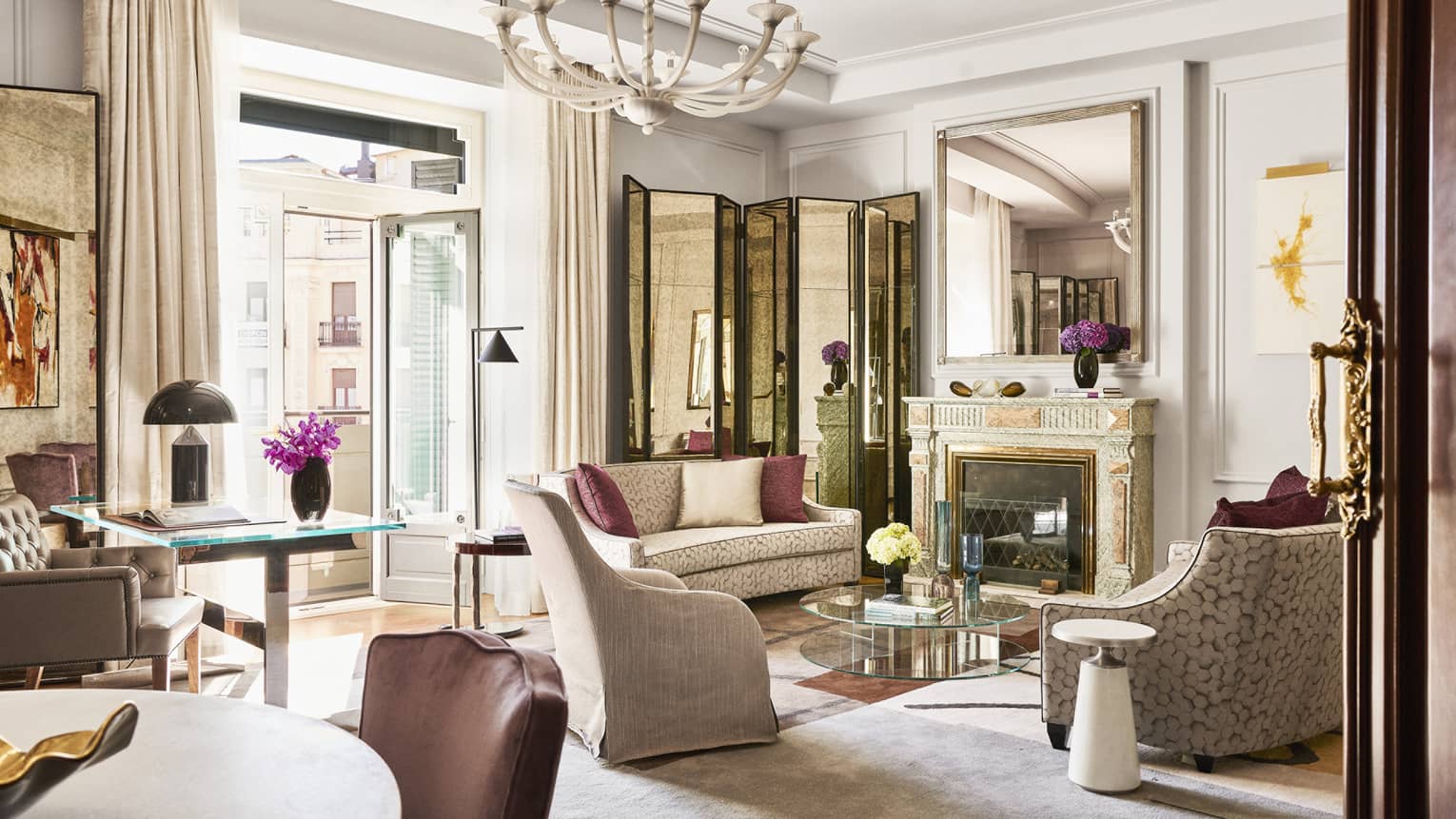 Luxury hotel suite with upholstered seating, glass coffee table, desk, dining table, decorative fireplace, large mirror, chandelier and open doors to balcony