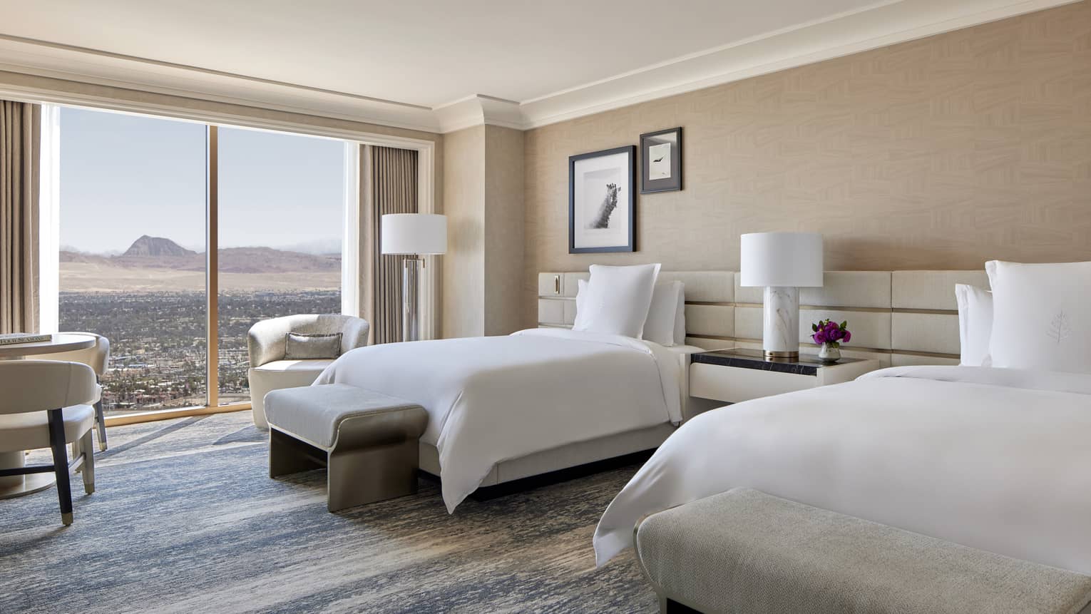 A room with two beds, large window, side tables, and chairs.