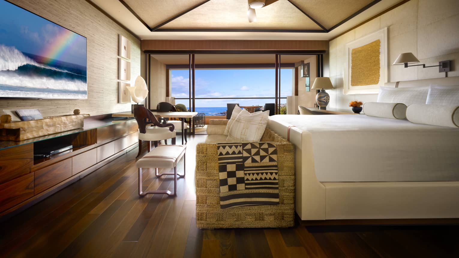Hotel suite bed and sofa at foot facing TV, seating area with floor to ceiling window and ocean view