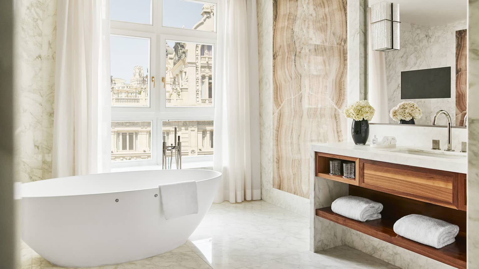 Luxury hotel bathroom with a freestanding bathtub, marble walls, vanity with wooden shelving, mirror, TV, large window with city view and elegant decor