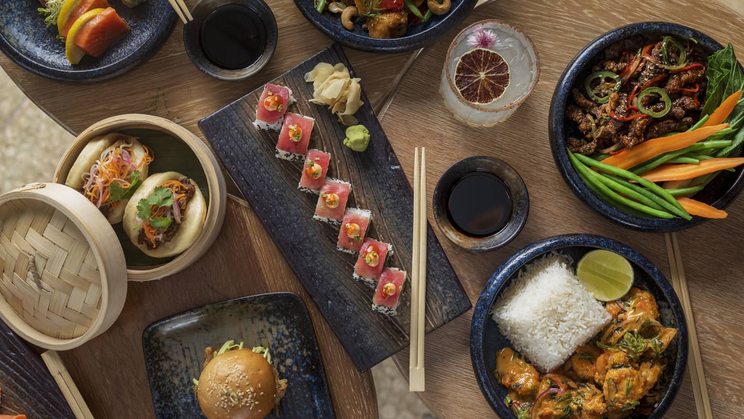 Wooden table filled with black plates of sushi, bao buns, shrimp and rice, and other assorted dishes