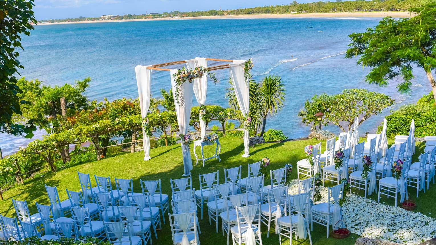 Aerial view of rows of white chairs facing wedding altar on lawn by ocean