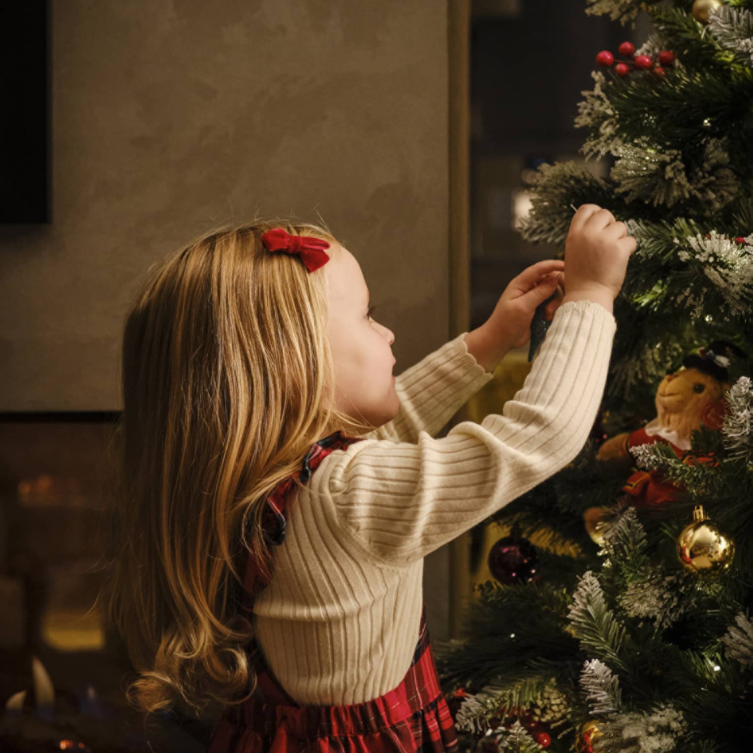 Close-up of a young child, red velvet barrette in their smooth blonde hair, arms reaching up to decorate a Christmas tree.