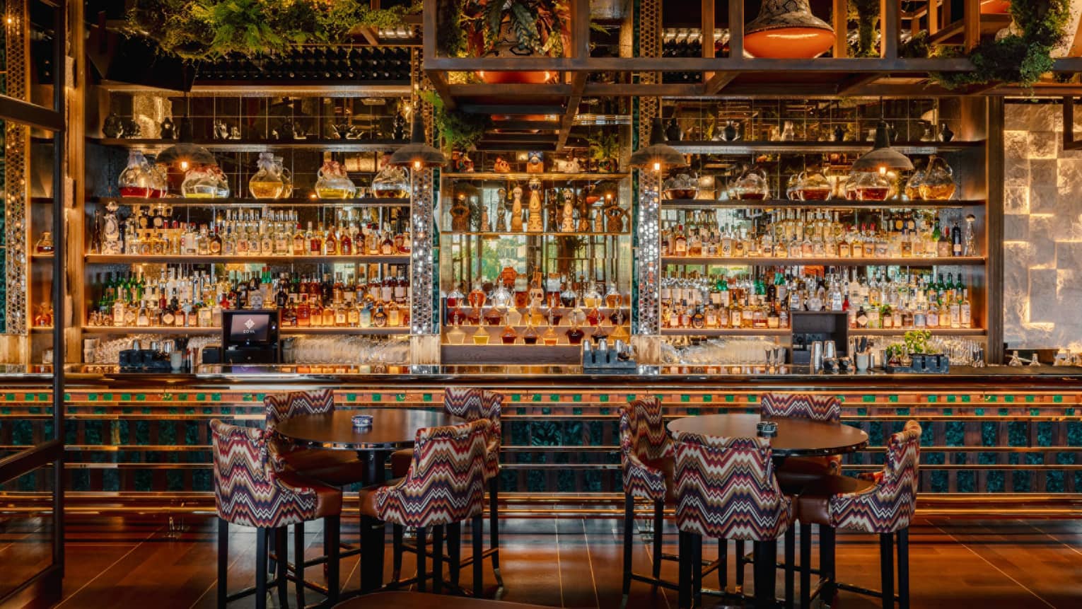 Two cocktail tables sit in front of a tiled bar counter stocked with four rows of glass shelves filled with bottles