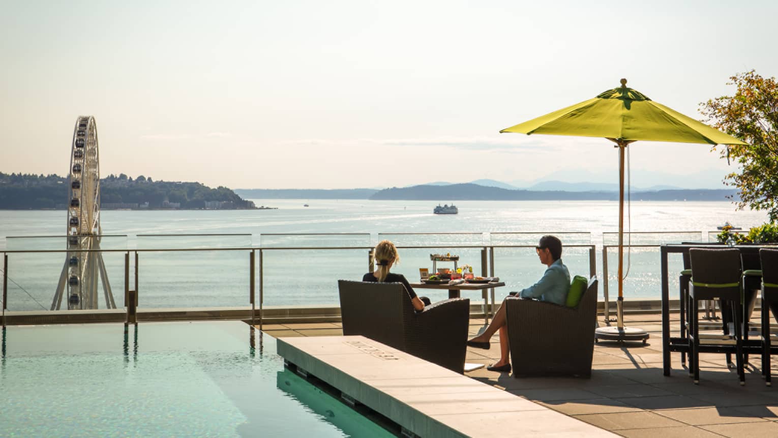 Couple sits poolside at dining table on sunny patio overlooking Elliott Bay and The Seattle Great Wheel
