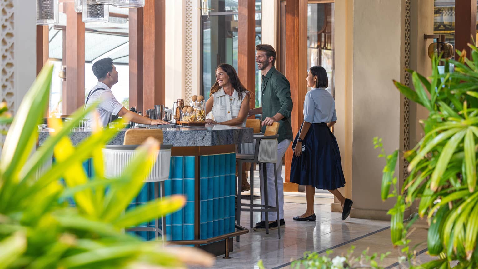 Three people walk up to an open-air, blue-tiled bar surrounded by green plants