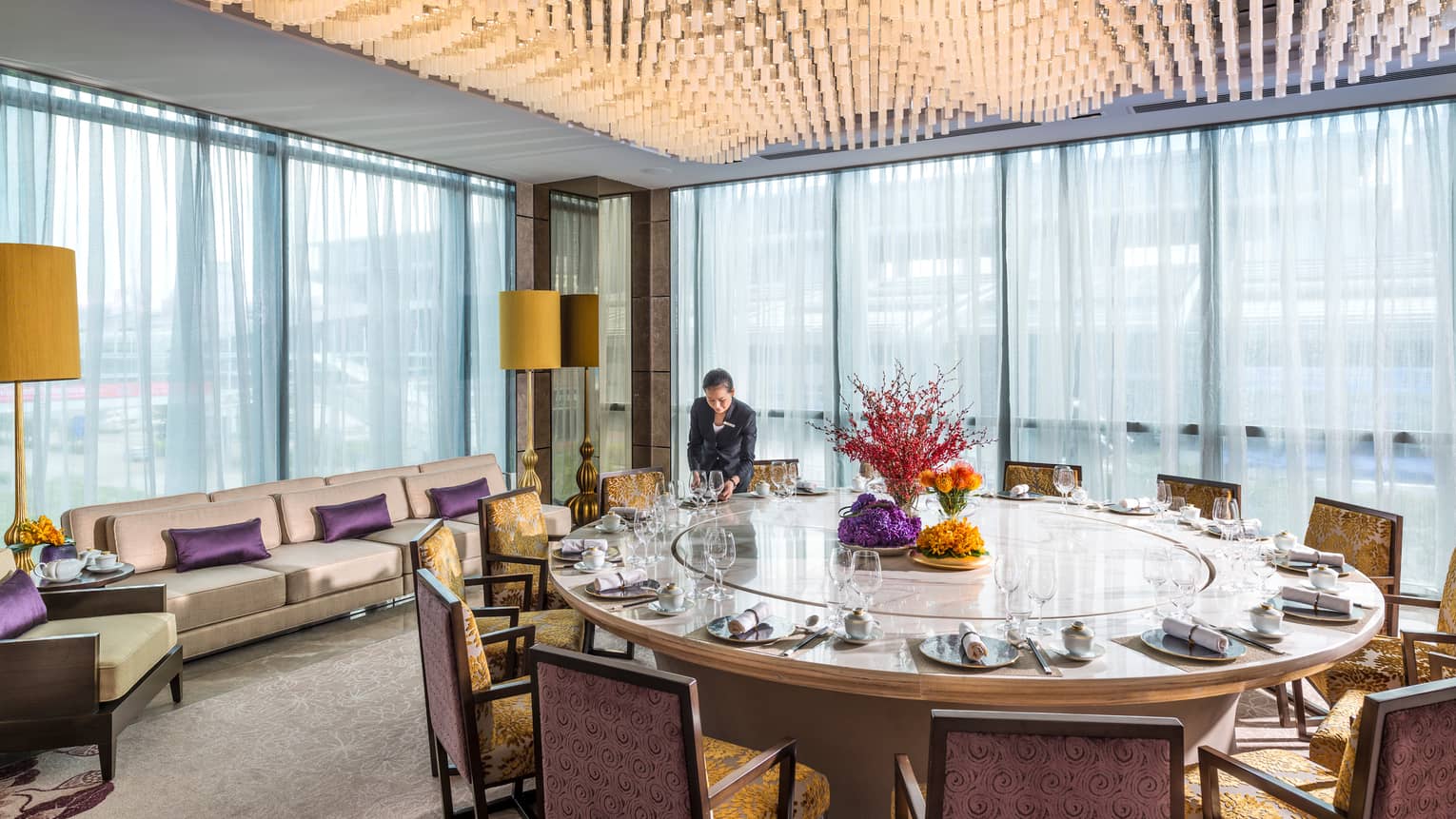 Server sets large round dining table by corner windows under ceiling covered with hanging lights