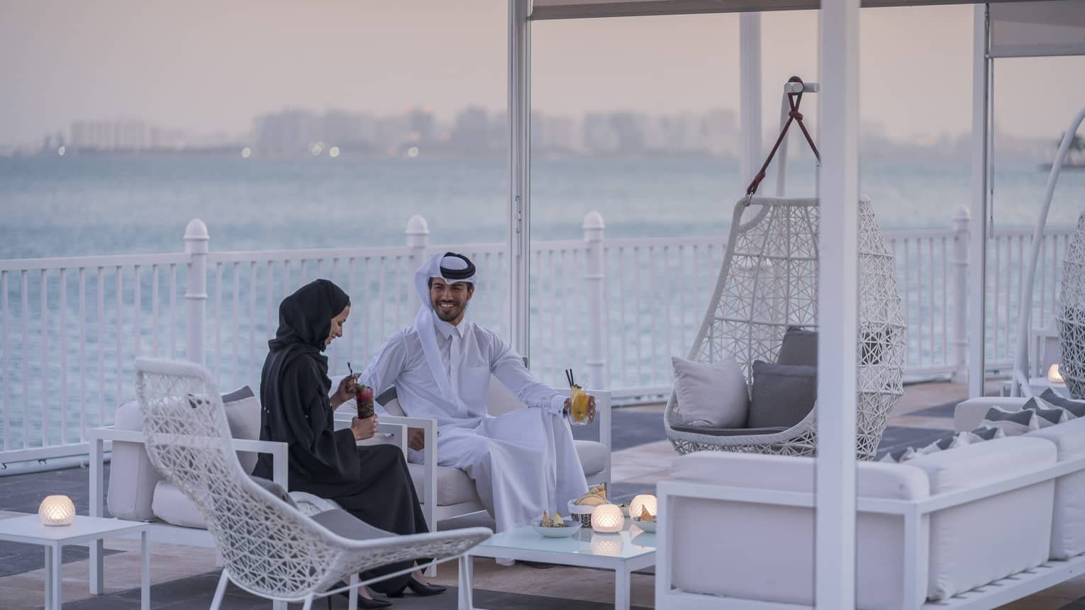 Man and woman sit on sofa on outdoor, covered terrace next to the water