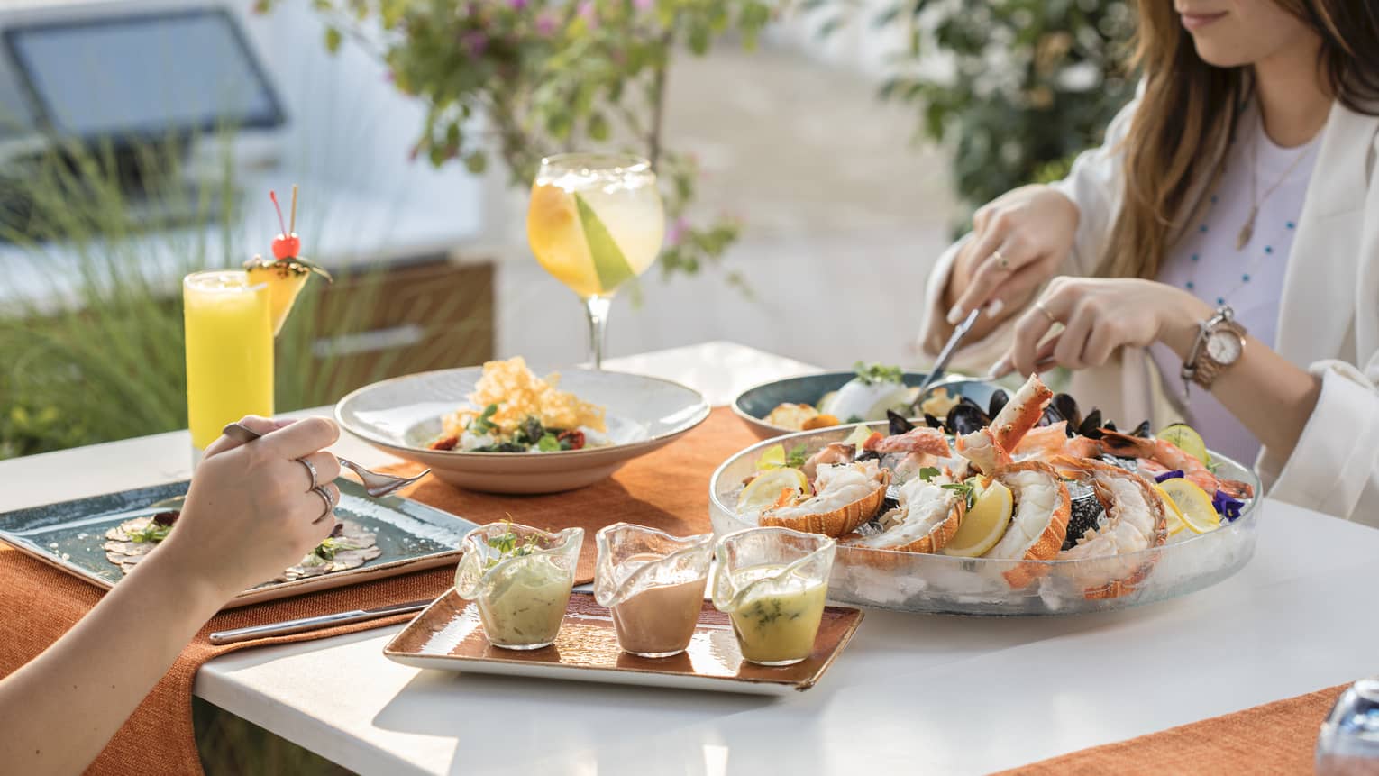 Two people sit at an outdoor table set with a large seafood platter on ice, a plate of three sauces and three other plates of food along with two tropical beverages
