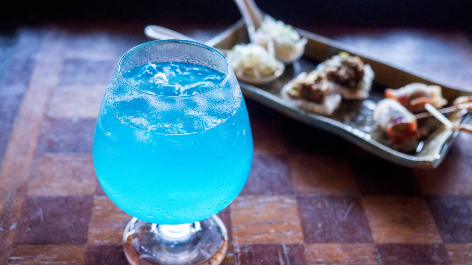 Bright blue cocktail in glass beside platter with small appetizers