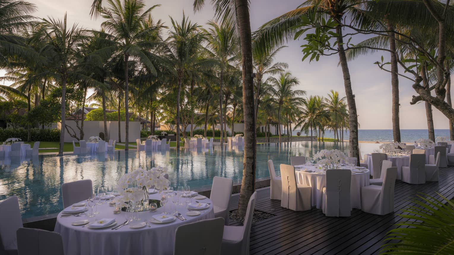 Round dining tables with white tablecloths, white chairs beside infinity pool and palm trees
