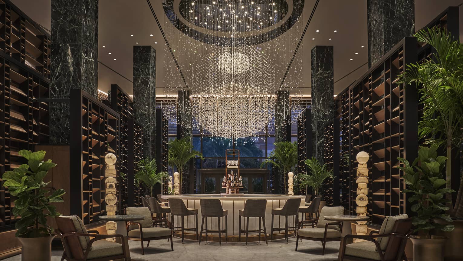 Chandelier Bar with six jade pillars flanked by wooden shelves and potted palms, glass-topped bar in the middle with six cocktail chairs, giant chandelier hanging overhead