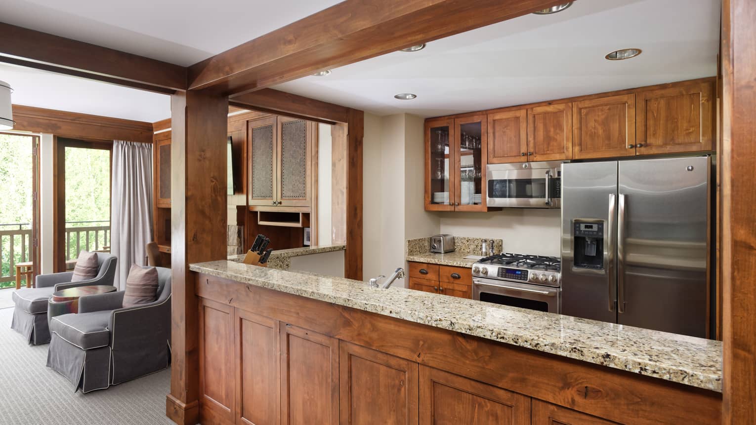 Kitchen with wooden cabinets, stainless steel appliances and granite countertops, opening to a living room with two grey armchairs
