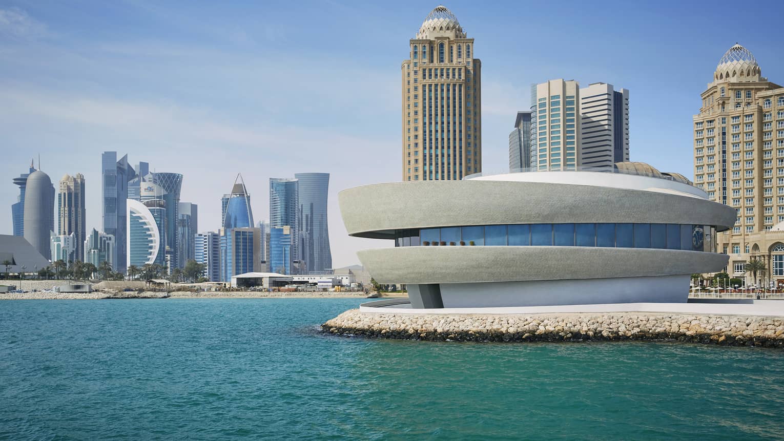 Exterior view of Nobu Doha, a round building with windows in the center sitting on the edge of the water with the skyline behind it