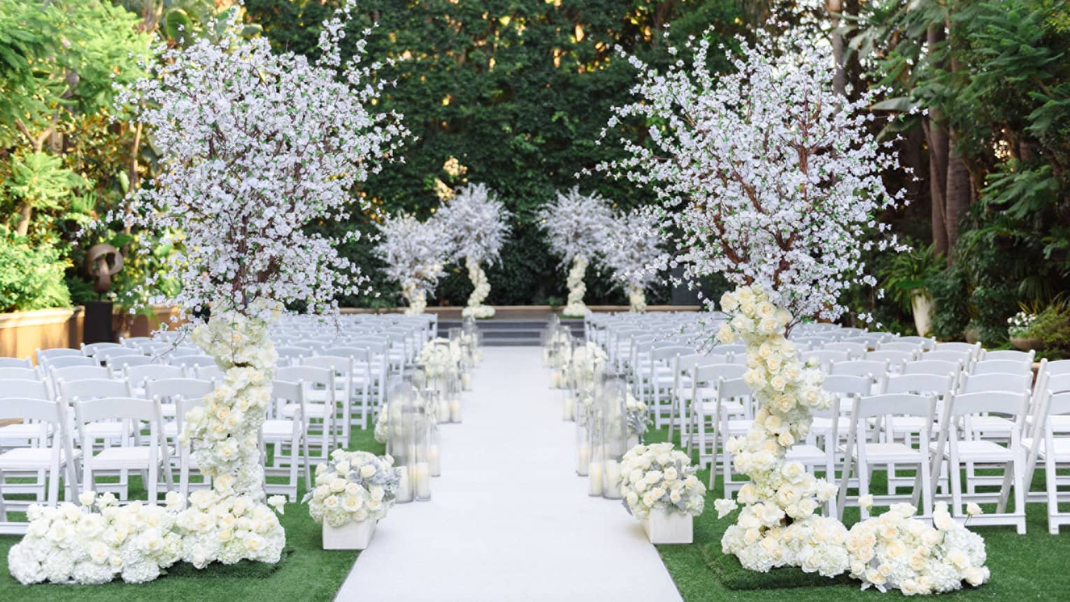 An outdoor wedding event space with rows of white chairs, white floral decor that includes flowering trees, and a white walkway leading to a raised platform in the background. The area is surrounded by trees and other greenery.