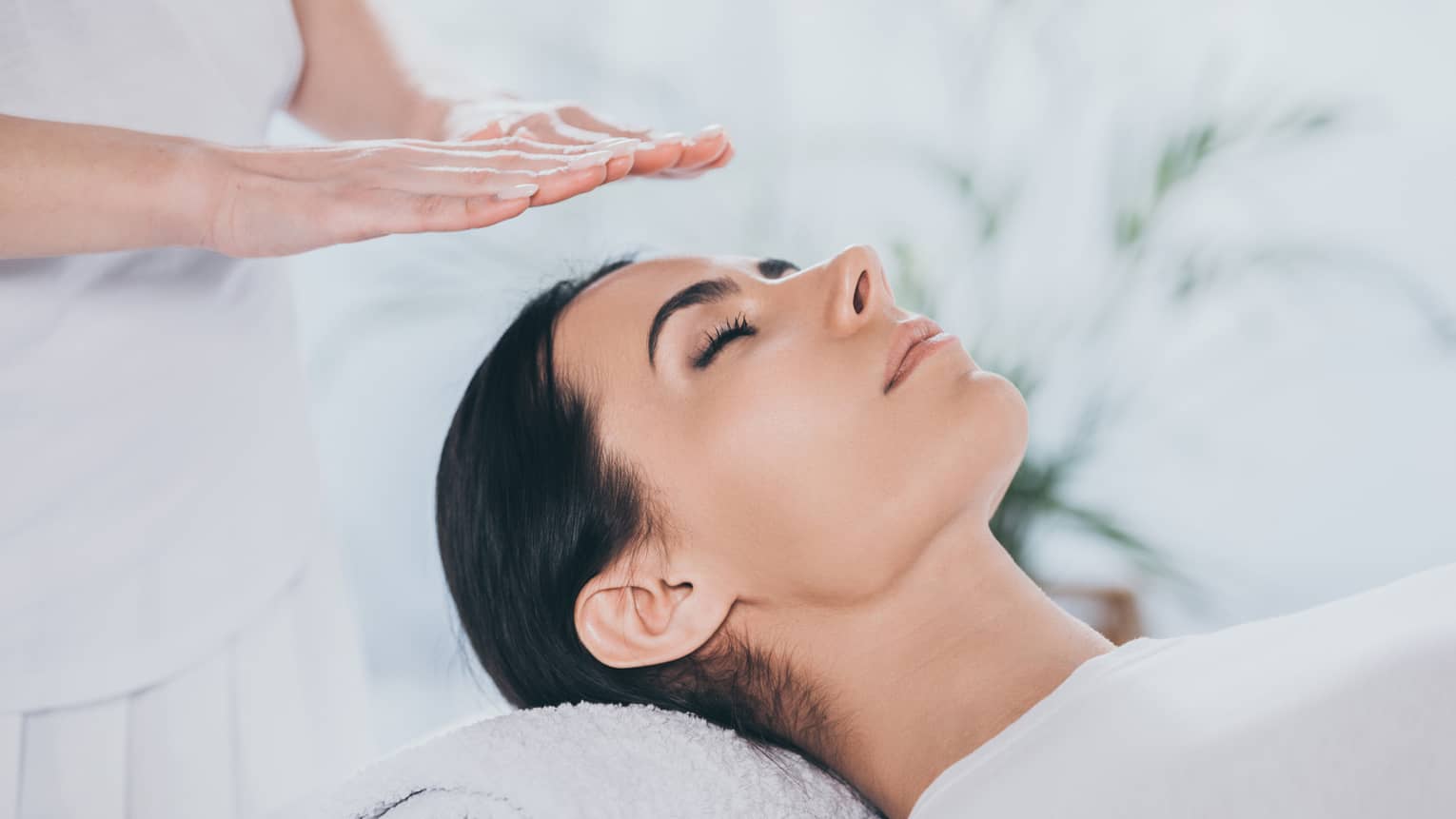 Woman with eyes closed lays on rolled up towel on spa table under therapists' hovering hands